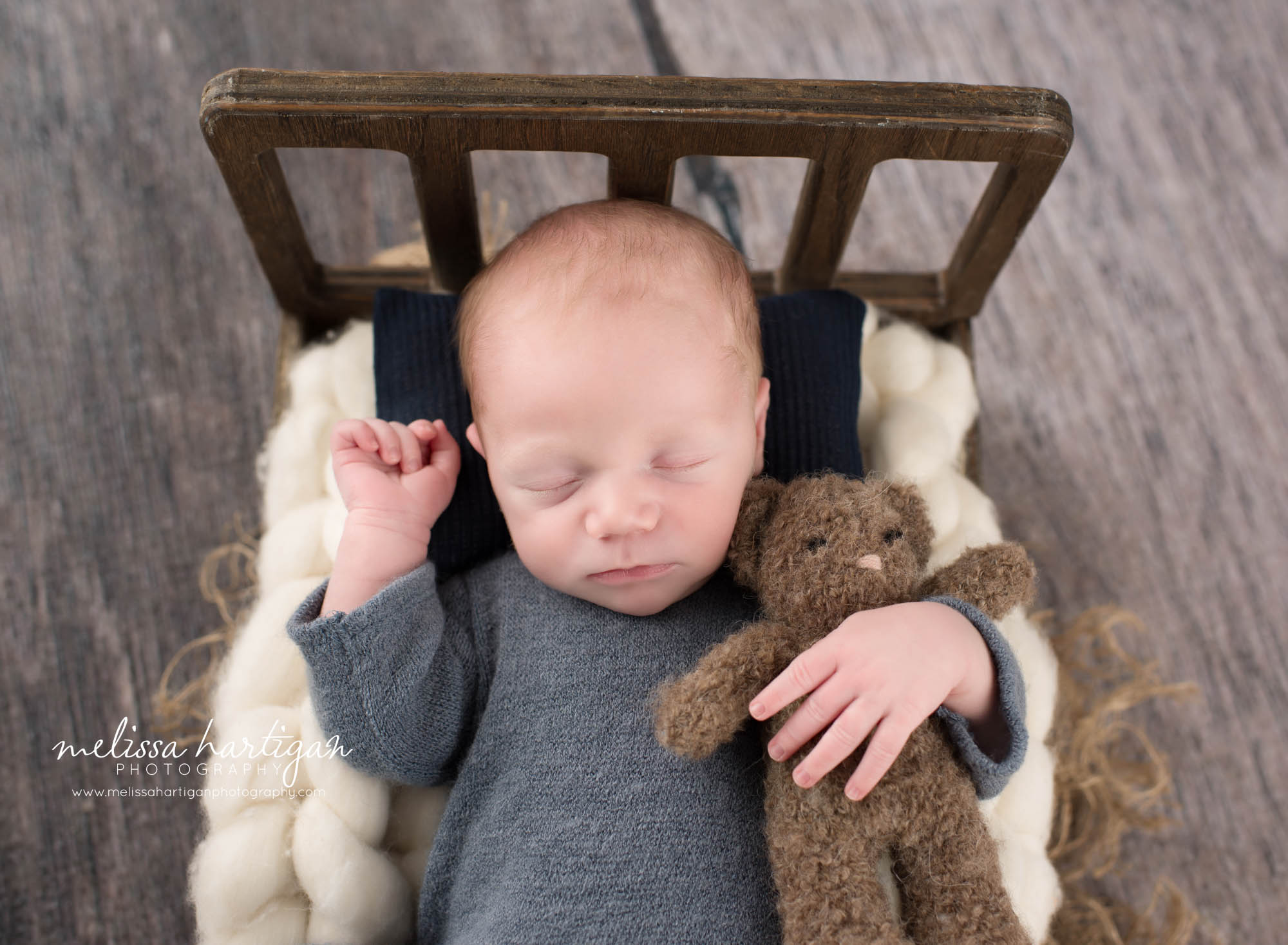 newborn baby boy posed on back on wooden bed prop holding brown teddy bear