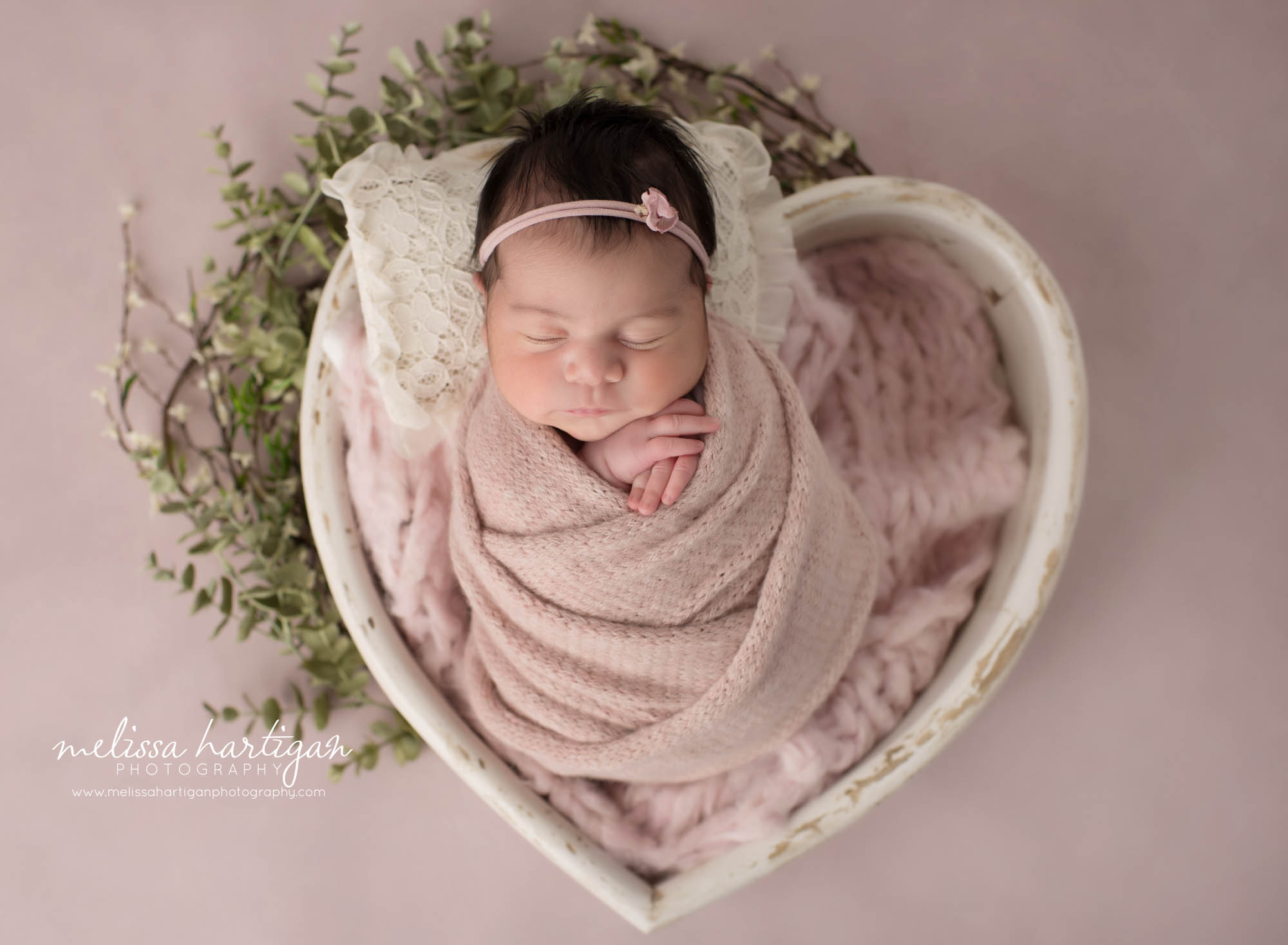 newborn baby girl posed in cream heart shaped wooden prop swaddled in pink knitted wrap Windham Baby Photography