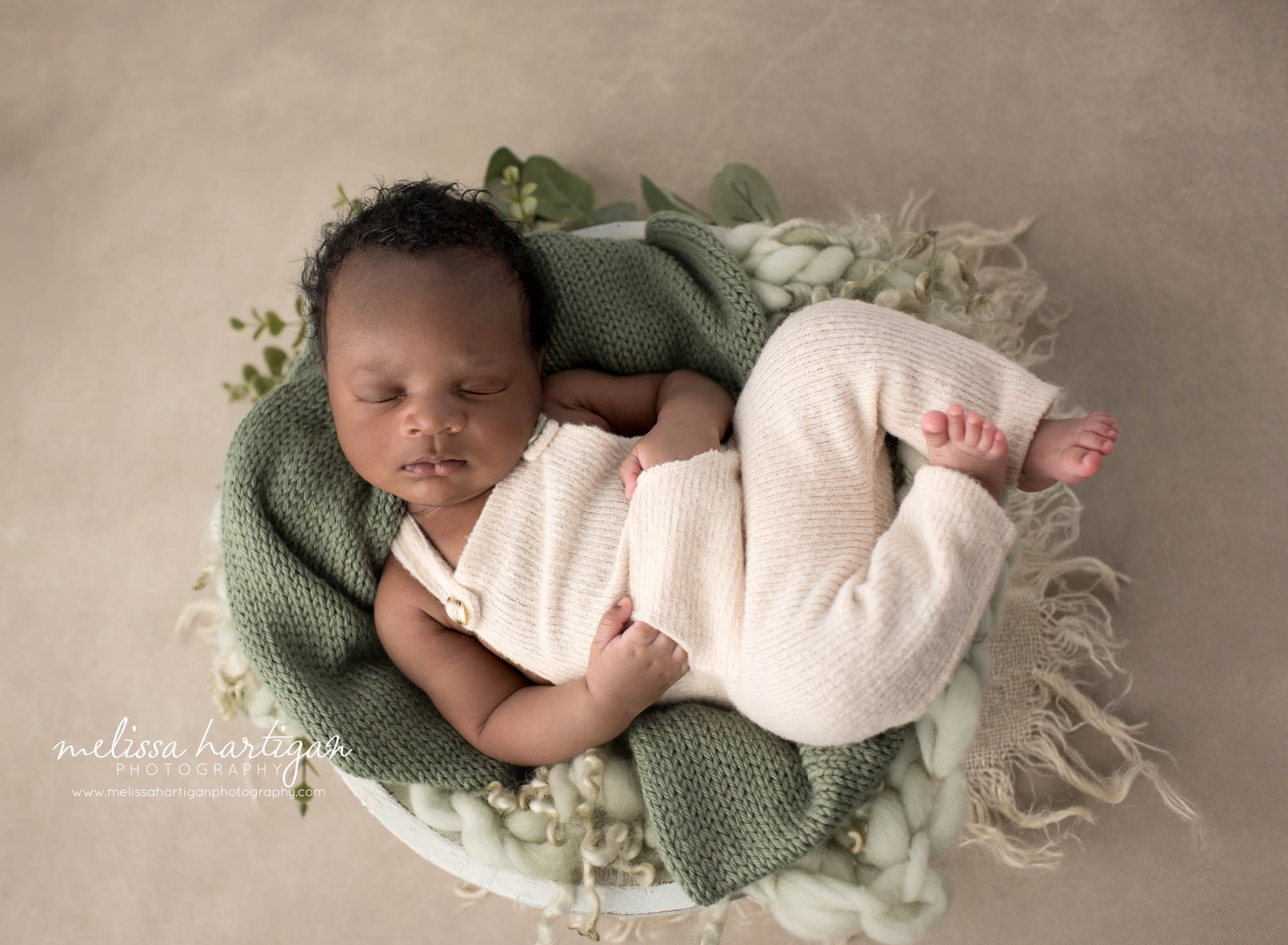 newborn baby boy posed in bowl prop with green knitted layer wrap wearing cream outfit with hands in pockets Newborn photography Hartford County