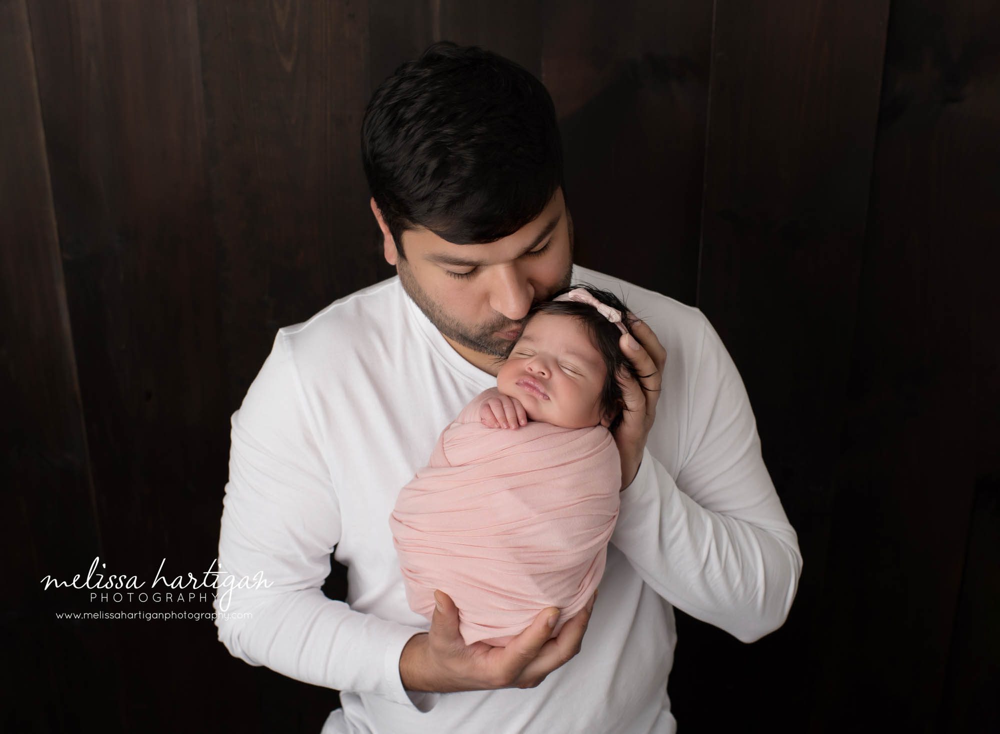 dad holding newborn daughter swaddled in pink wrap kissing her on side of forehead