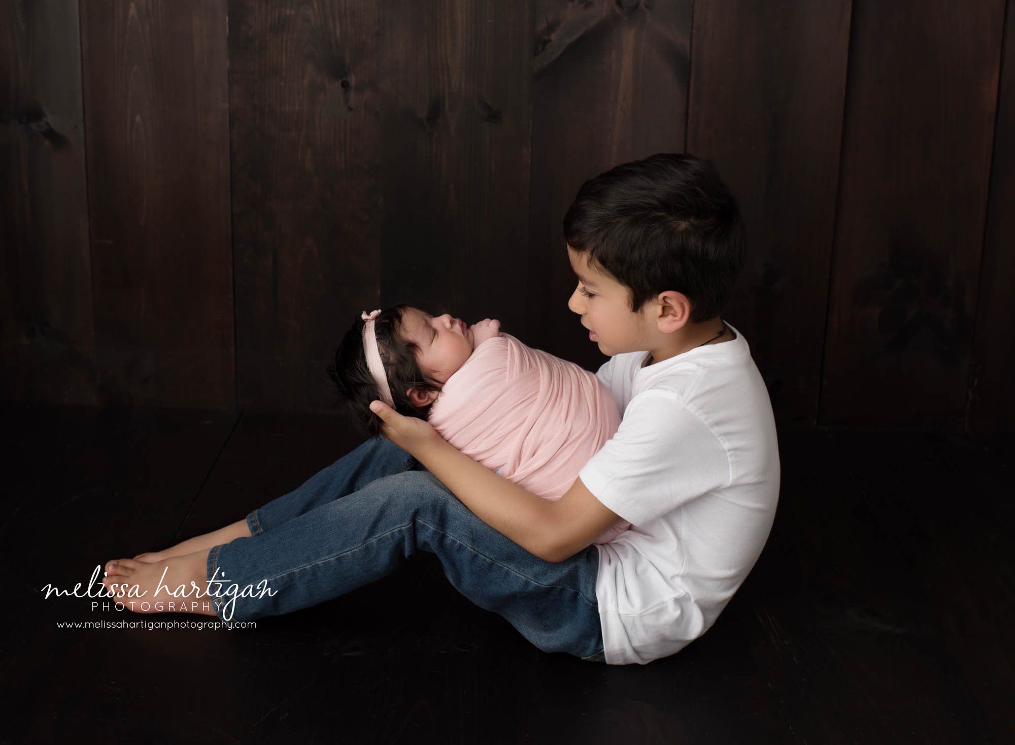 big brother sitting on floor sideways holding his new baby sister swaddled in pink wrap Suffield CT Newborn photography