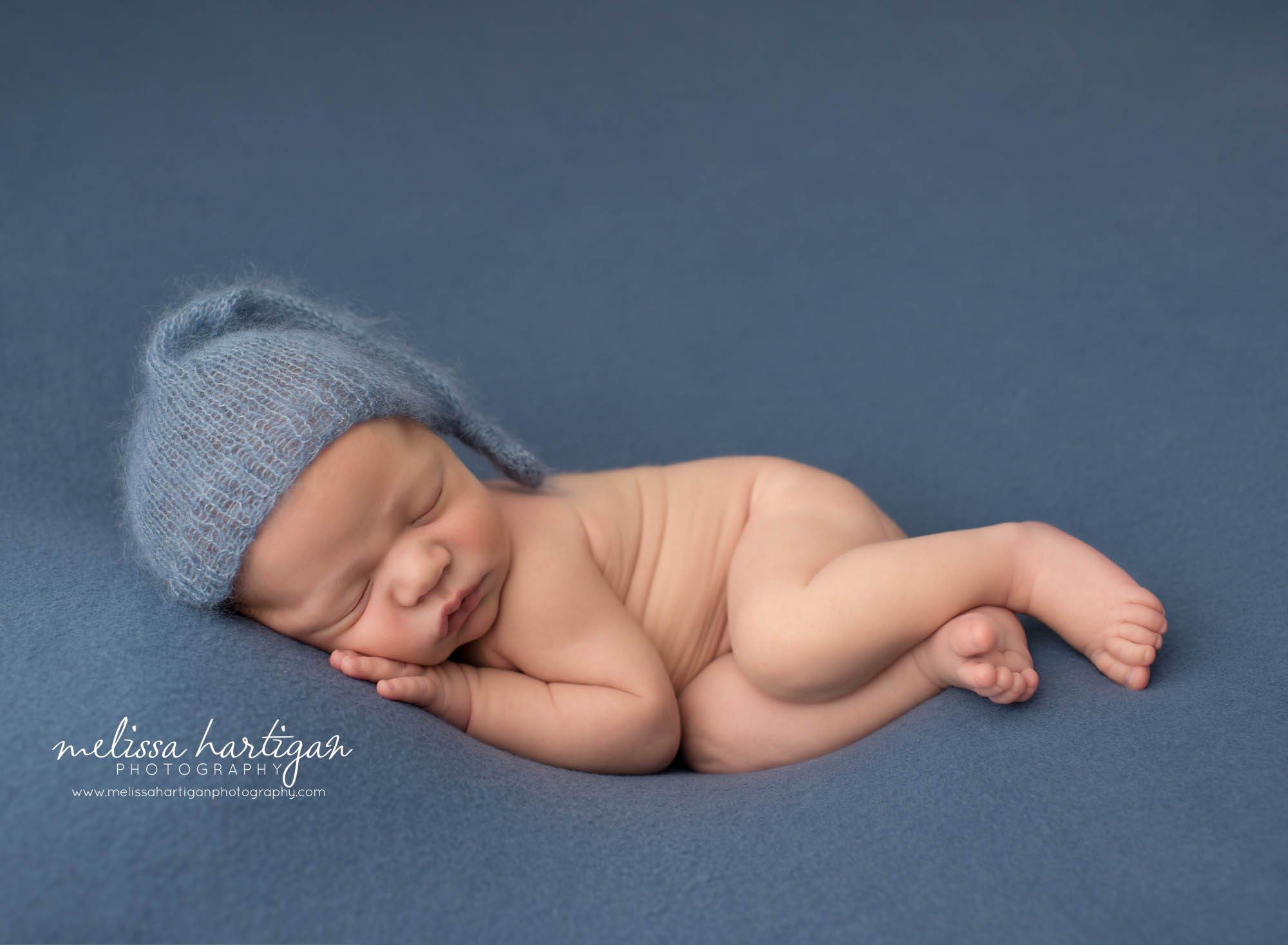newborn baby boy posed on b lue backdrop wearing knitted sleepy cap