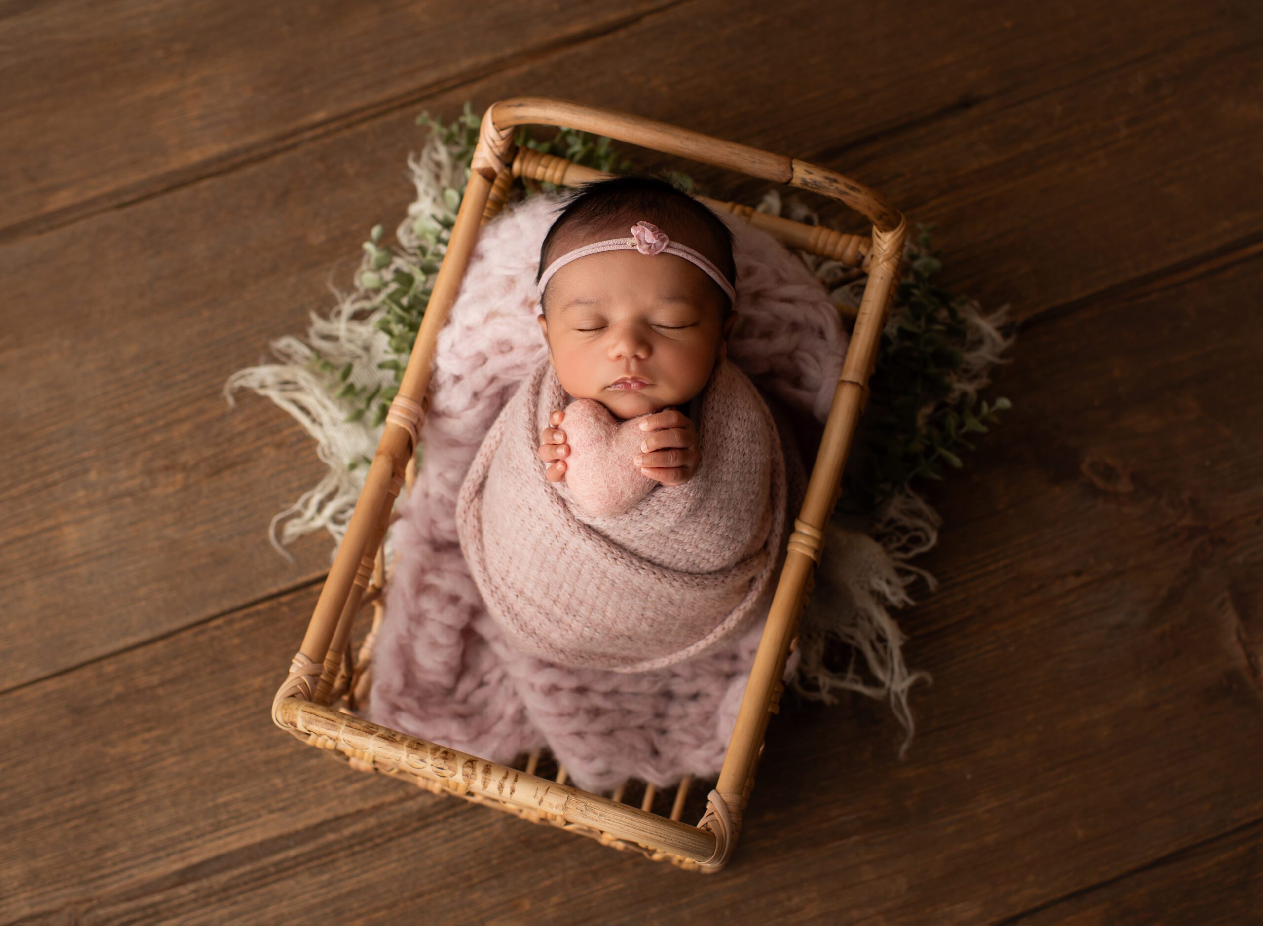 Newborn baby girl posed in basket holding hearted felt prop wrapped in pink wrap