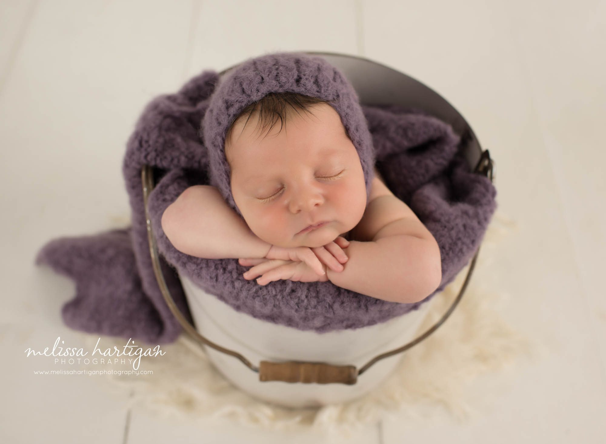 Newborn baby girl posed in bucket wearing purple knitted bonnet