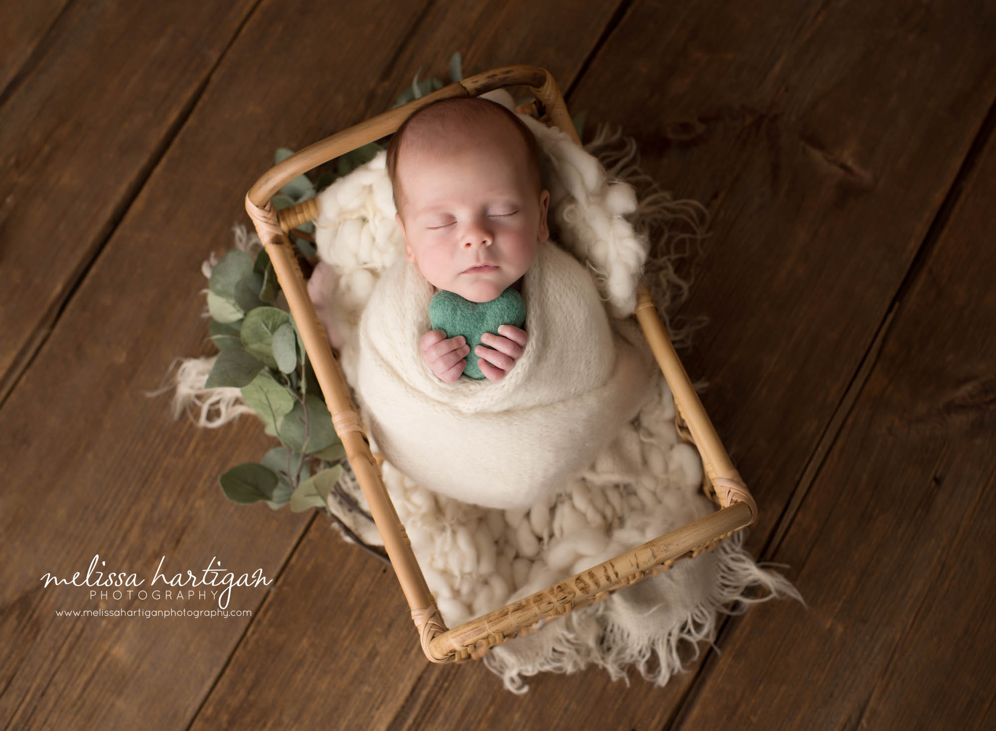 Newborn baby boy posed in basket swaddled in knitted cream wrap holding green felted heart prop
