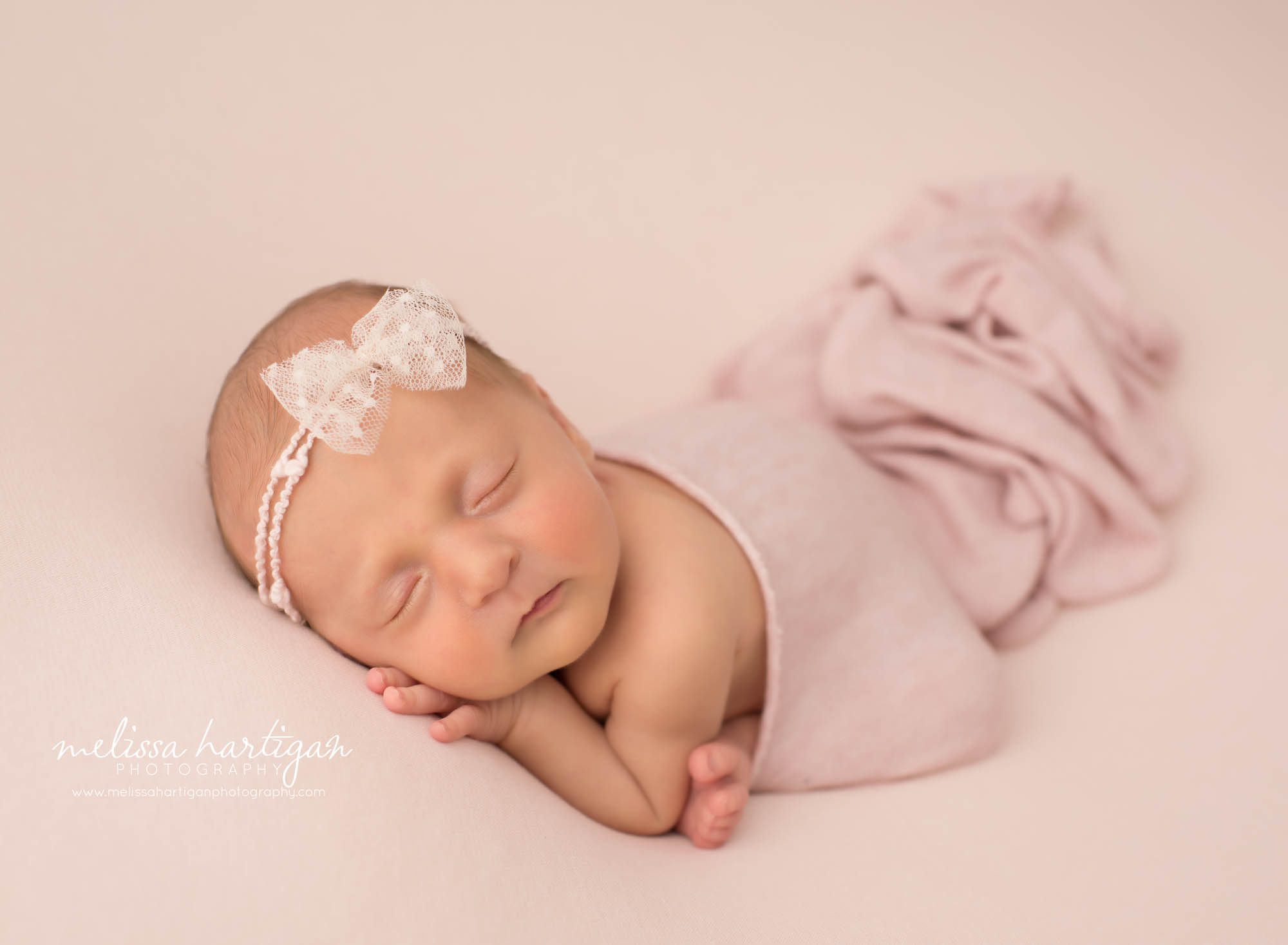 newborn baby girl posed on pink backdrop with pink layer draped over her wearing bow headband