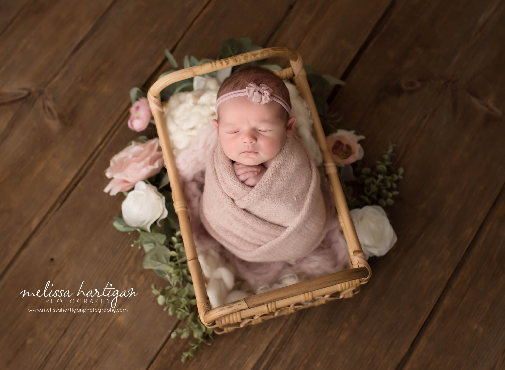 Newborn baby girl posed in basket wearing pink bow headband swaddled in knitted pink wrap