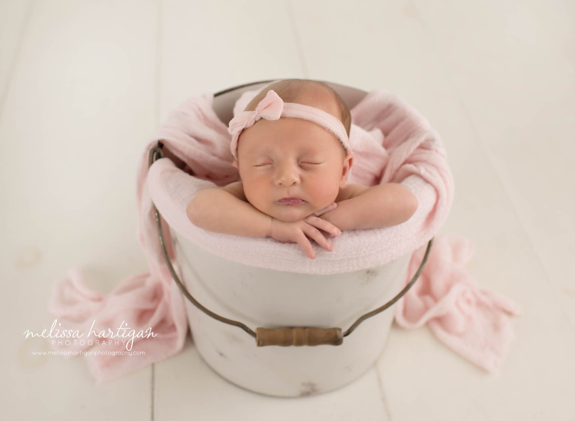 Newborn baby girl posed in bucket with pink layer draping over and wearing bow headband