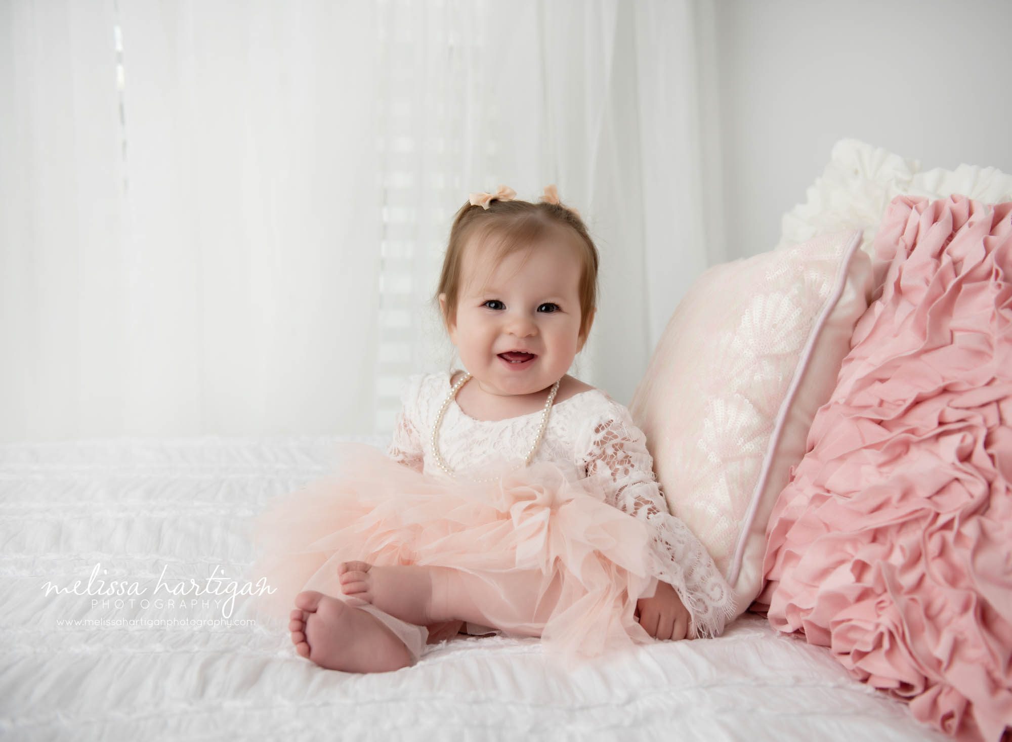 Baby girl sitting on bed in studio happy smiling wearing beads and pink and cream lace dress Preston CT Baby Photography