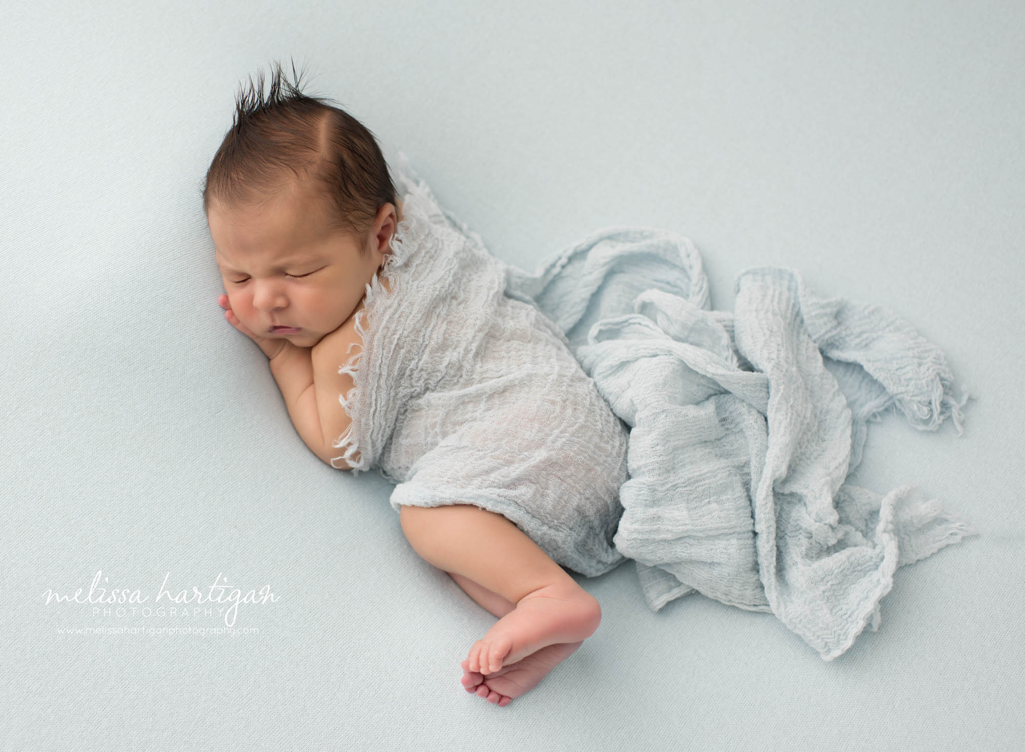 Newborn baby boy posed on side sleeping with hand under cheek draped in blue cheesecloth layer