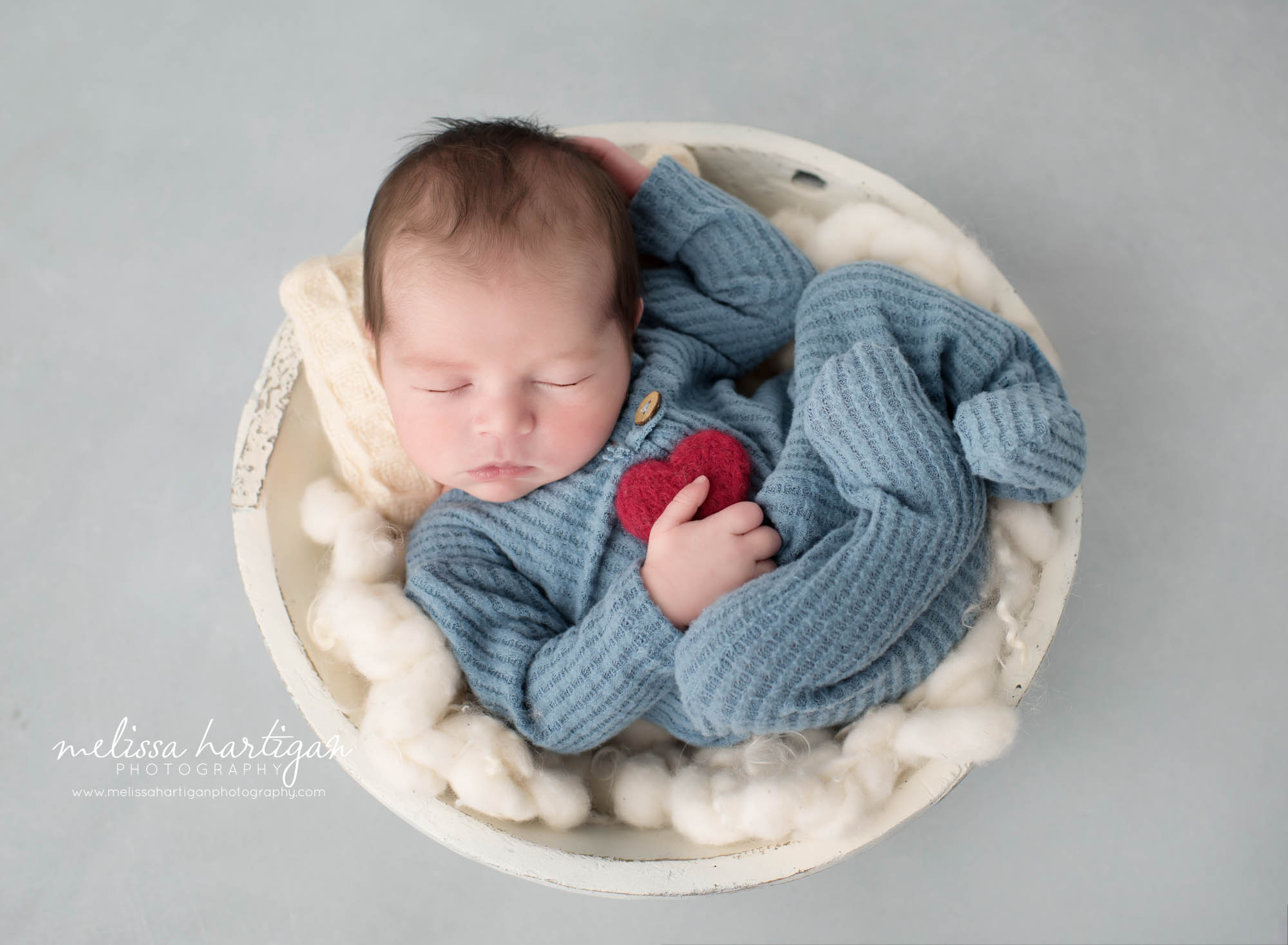 Newborn baby boy wearing blue waffle knit footed sleeper holding red felted heart prop posed in wooden bowl