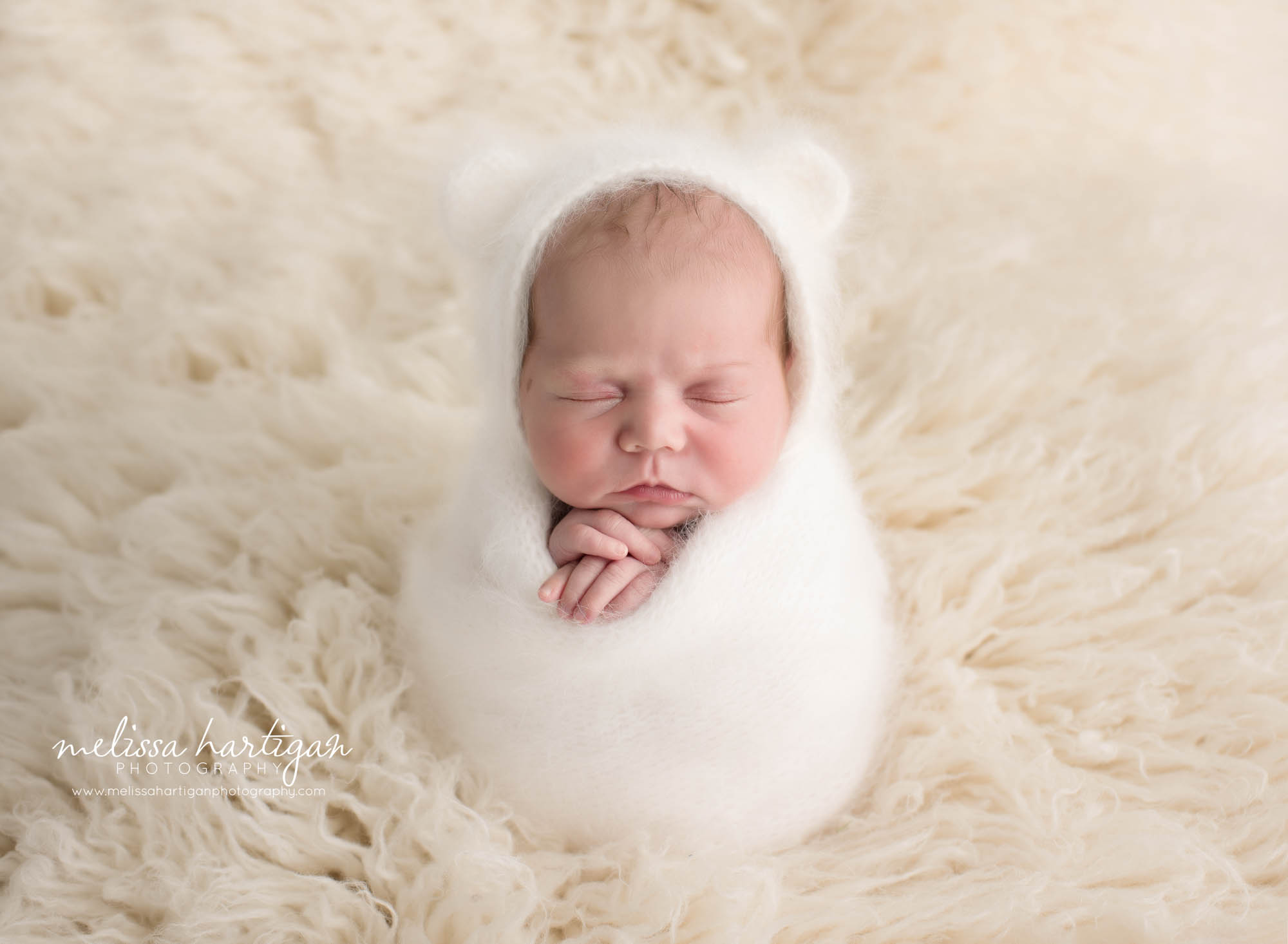 Newborn baby girl swaddled in wrap wearing knitted bear bonnet in white