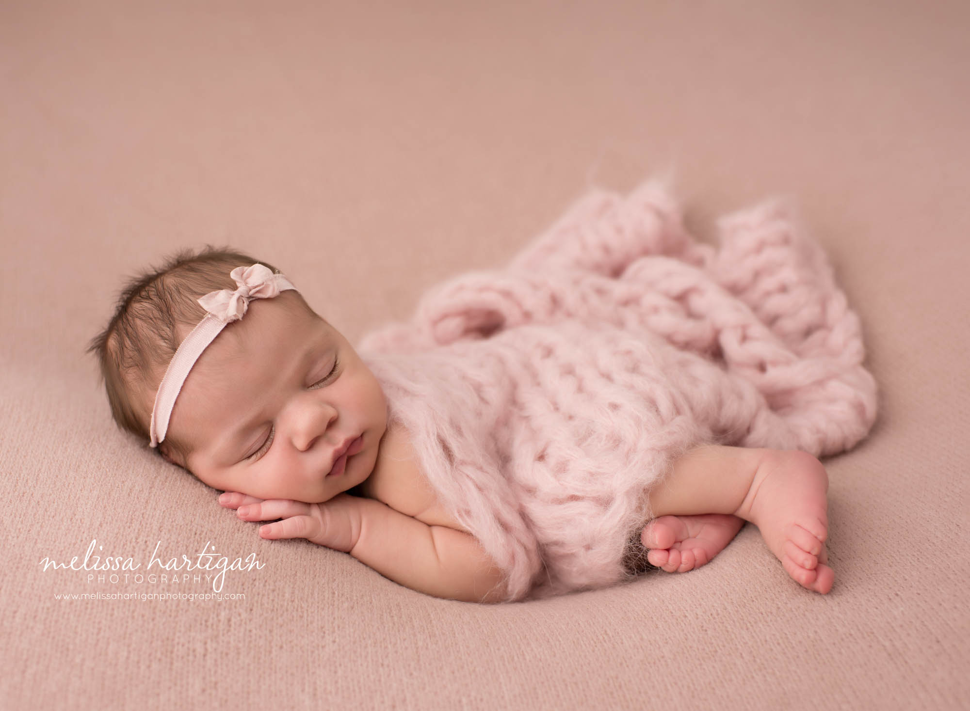 Newborn baby girl posed on pink backdrop wearing pink bow headband and pink knit layer draped over Newborn Photography Norwich CT