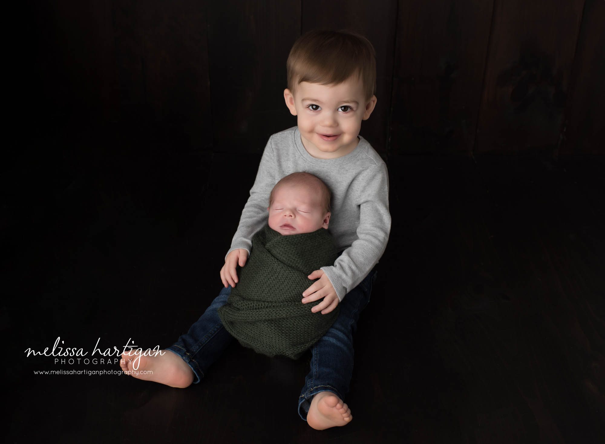 Big brother sitting in studio with newborn baby brother swaddled in green knit wrap Newborn Photography Vernon CT