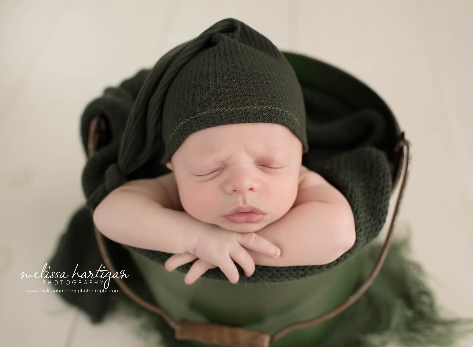 Newborn baby boy posed in green bucket wearing green sleepy cap