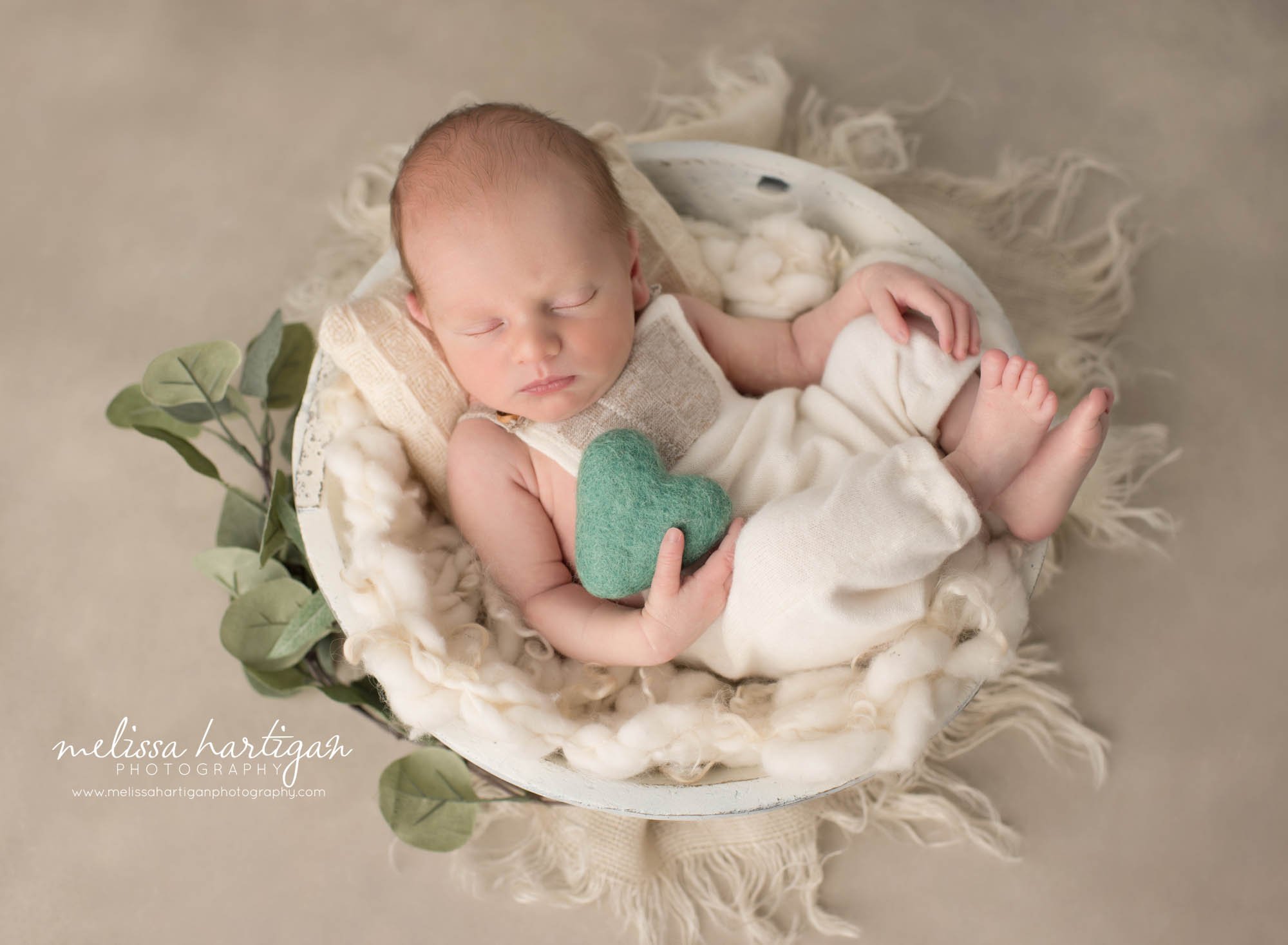 Newborn baby boy posed in wooden bowl with cream layer wearing cream outfit and holding green felted heart prop Newborn Photography Ledyard CT